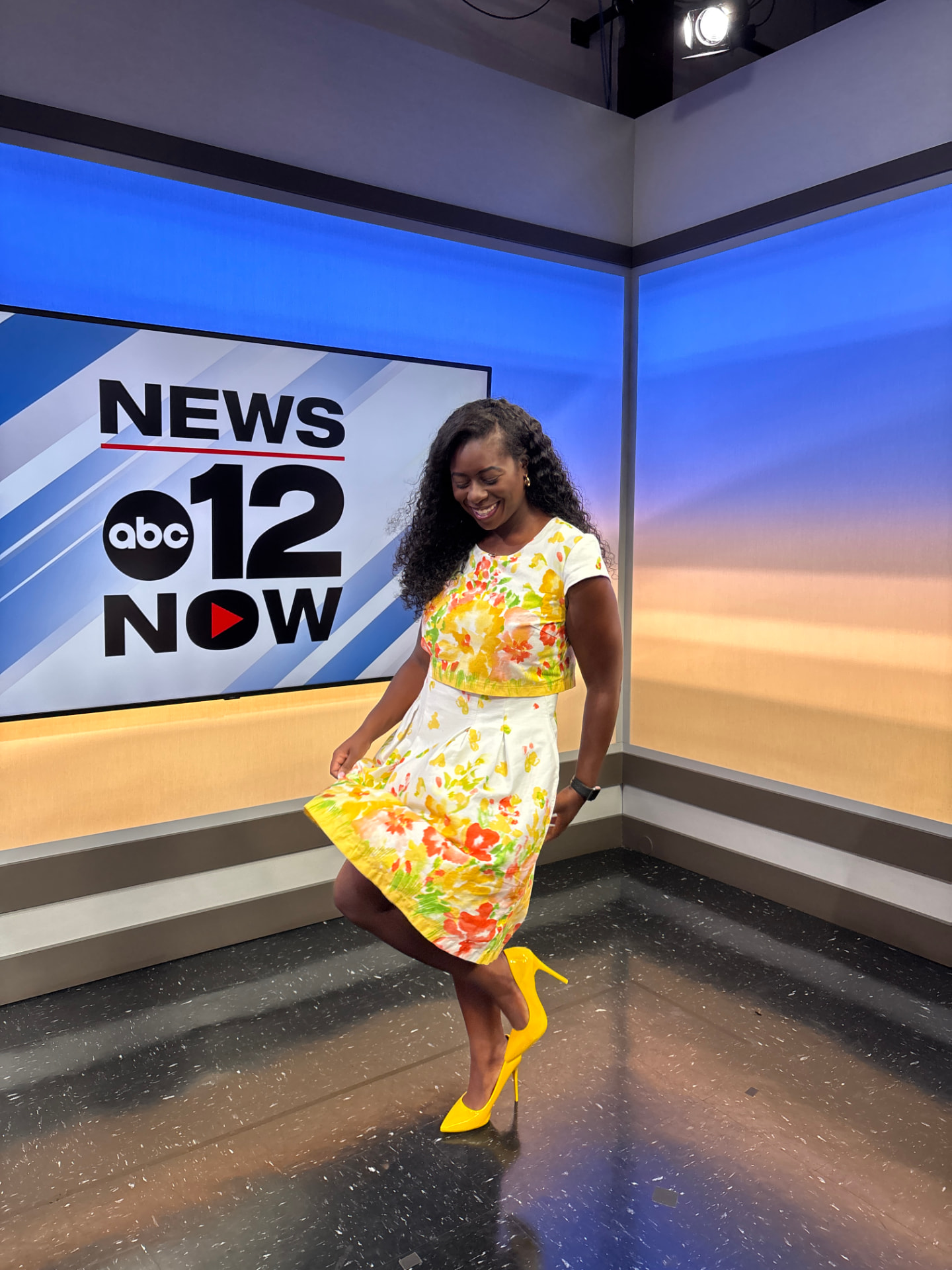 Moriah Davis laughing in a yellow floral dress and yellow heels on the news set