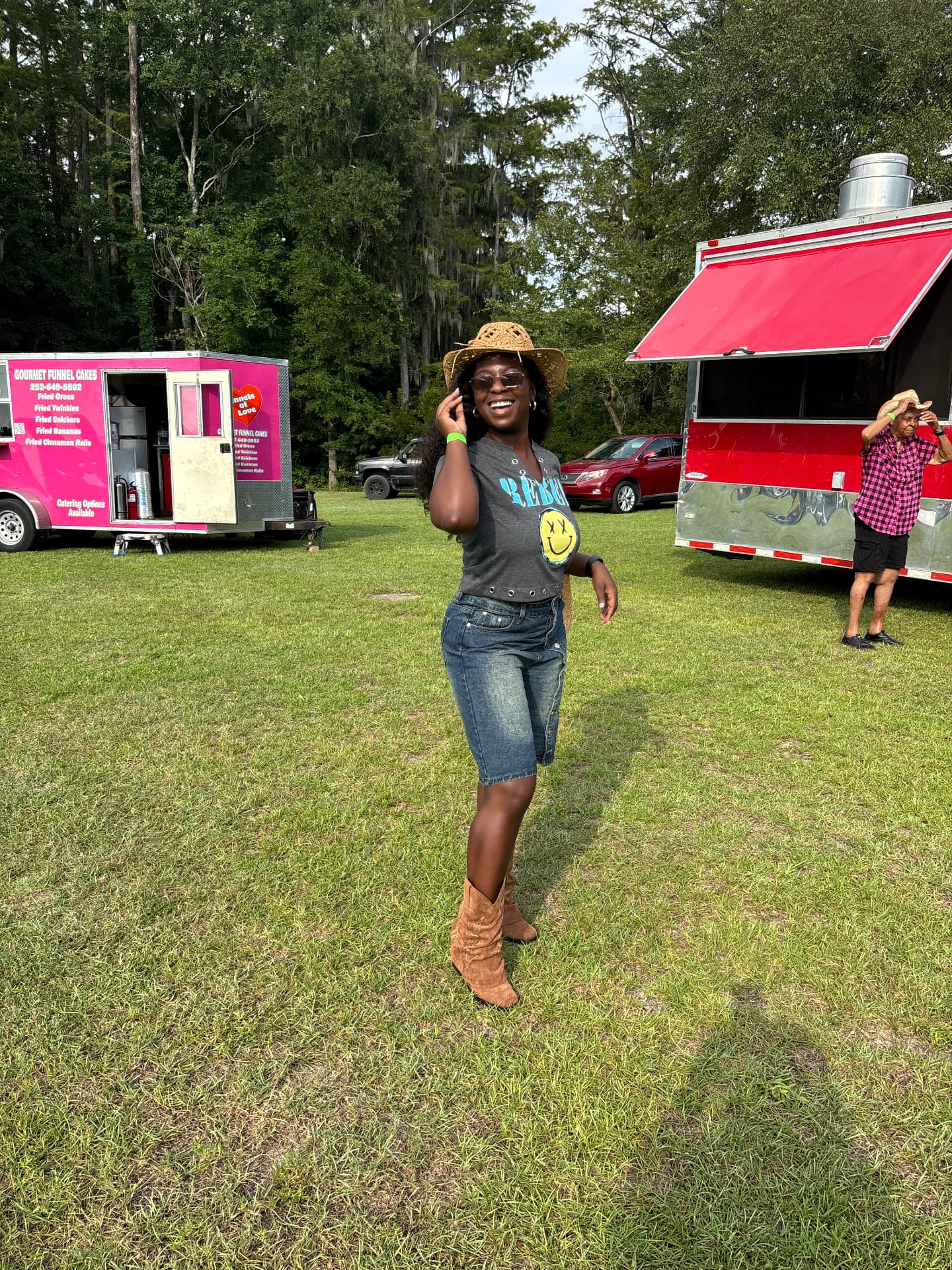 Moriah Davis smiling in a straw cowboy hat, smiley-face graphic tee, denim shorts and tan cowboy boots at an outdoor festival